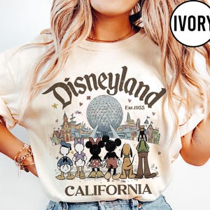 May include: Ivory-colored Disneyland California graphic t-shirt. The design features the Disneyland logo, the iconic Spaceship Earth, and cartoon characters like Mickey and Minnie Mouse. The text "Est.1955" is also included.