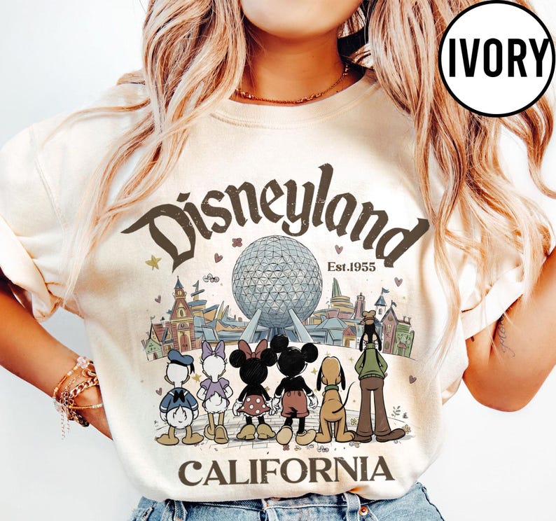 May include: Ivory-colored Disneyland California graphic t-shirt. The design features the Disneyland logo, the iconic Spaceship Earth, and cartoon characters like Mickey and Minnie Mouse. The text "Est.1955" is also included.