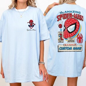 K&ouml;nnte beinhalten: Hellblaues T-Shirt mit Spider-Man-Motiven. Die Vorderseite zeigt ein kleines Spider-Man-Logo und Text. Die R&uuml;ckseite zeigt ein gro&szlig;es Spider-Man-Maskendesign mit dem Text "The Amazing Spider-Man" und einen Bereich f&uuml;r einen individuellen Namen.