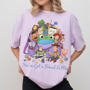 May include: A light purple t-shirt featuring a graphic print of various animated characters from Toy Story emerging from a purple backpack, with the text "You're Got a Friend In Me" printed in blue script lettering at the bottom of the design.