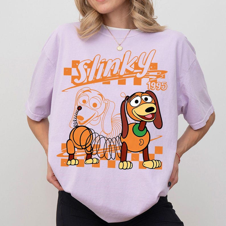 May include: A light purple short-sleeve t-shirt featuring a graphic of a Slinky Dog character with the text Slinky 1995. The design includes an orange checkered pattern background and a line-art outline of the dog alongside a colored version.