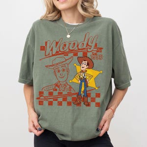 May include: An olive green t-shirt featuring a graphic print of the character Woody from Toy Story. The design includes the text Woody 1995, a red checkered pattern, a yellow sheriff star, and a line art illustration of the character's face.