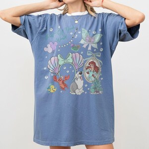 May include: A blue short-sleeve t-shirt featuring a graphic print with the text Part Of Your World, a mermaid character, a crab, a fish, a dog, a seashell headband, a bow, and bubbles. The design includes pastel colors and decorative elements.