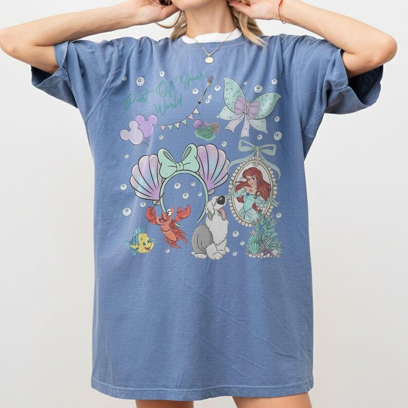 May include: A blue short-sleeve t-shirt featuring a graphic print with the text Part Of Your World, a mermaid character, a crab, a fish, a dog, a seashell headband, a bow, and bubbles. The design includes pastel colors and decorative elements.