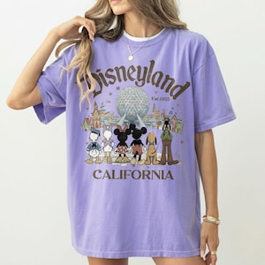 May include: A lilac-colored t-shirt with a Disneyland graphic. The graphic features the Disneyland logo, the text "Est.1955", and cartoon characters like Mickey and Minnie Mouse. The word "CALIFORNIA" is printed at the bottom of the graphic.