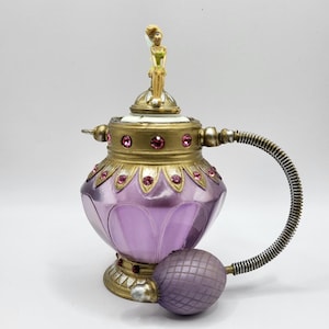 May include: A decorative perfume bottle with a purple glass body and gold accents. The bottle features a small fairy figurine on top and pink jewel embellishments. A bulb atomizer is attached to the side.
