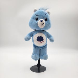 May include: A light blue plush bear with a grumpy expression, featuring a dark blue cloud and red heart design on its belly. The bear has a tuft of blue hair and is mounted on a black stand.