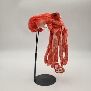 May include: A plush, coral-colored octopus hat with large, round eyes and long tentacles. The hat has a silver-colored mouth area and a red and gold tag. The hat is displayed on a black stand.