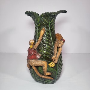 May include: A decorative vase shaped like a large green leaf, with two monkeys climbing on it. One monkey wears a red vest and the other a fez. The monkeys are tan with brown fur. The vase is a unique home decor item.