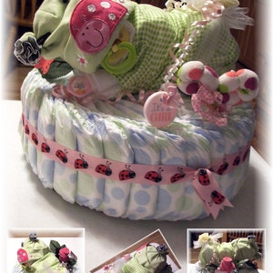 K&ouml;nnte beinhalten: Eine Windeltorte, verziert mit einem gr&uuml;n-wei&szlig; karierten Baby-Outfit, einem Marienk&auml;ferband und einem rosa Schild mit der Aufschrift "It's a Girl". Die Torte ist von rosa und wei&szlig;en Blumen umgeben.