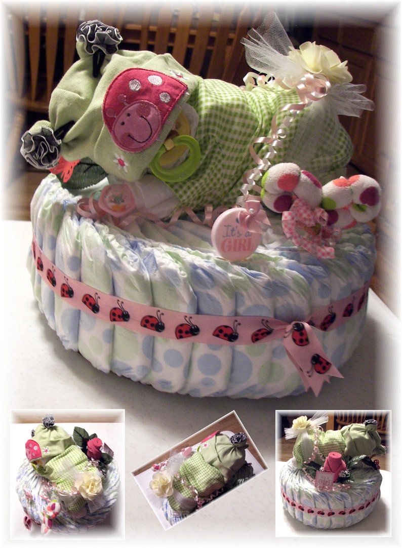 K&ouml;nnte beinhalten: Eine Windeltorte, verziert mit einem gr&uuml;n-wei&szlig; karierten Baby-Outfit, einem Marienk&auml;ferband und einem rosa Schild mit der Aufschrift "It's a Girl". Die Torte ist von rosa und wei&szlig;en Blumen umgeben.