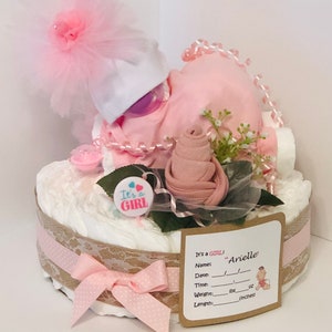 K&ouml;nnte beinhalten: Eine rosa und wei&szlig;e Windeltorte mit einer rosa Rose, einem wei&szlig;en Hut mit einer rosa T&uuml;llschleife und einer rosa und blauen "It's a Girl"-Nadel. Die Torte ist mit einem Jutestoffband und Spitze verziert. Eine braune Karte mit dem Text "It's a GIRL! Name: "Arielle" Date: Time: Weight:  lbs oz Length: inches" ist an der Torte befestigt.