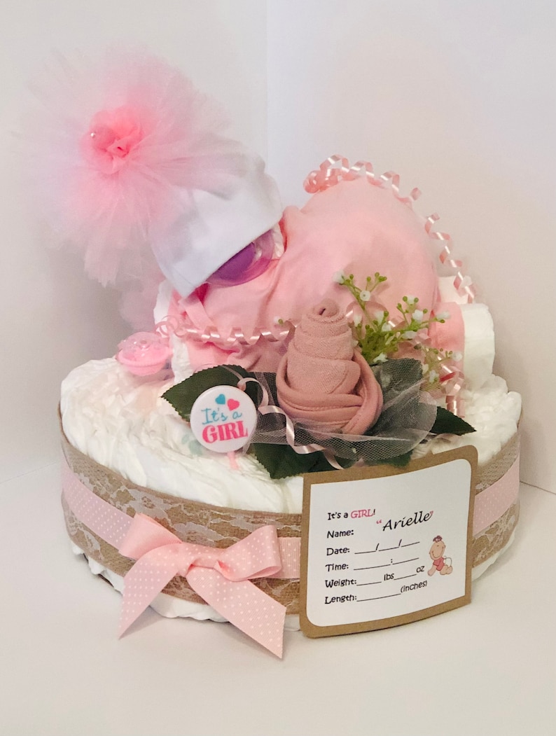 K&ouml;nnte beinhalten: Eine rosa und wei&szlig;e Windeltorte mit einer rosa Rose, einem wei&szlig;en Hut mit einer rosa T&uuml;llschleife und einer rosa und blauen "It's a Girl"-Nadel. Die Torte ist mit einem Jutestoffband und Spitze verziert. Eine braune Karte mit dem Text "It's a GIRL! Name: "Arielle" Date: Time: Weight:  lbs oz Length: inches" ist an der Torte befestigt.