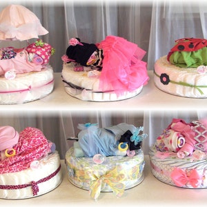 K&ouml;nnte beinhalten: Sechs Windelkuchen, dekoriert mit Babykleidung, B&auml;ndern und anderen babybezogenen Dekorationen. Die Kuchen sind in zwei Reihen zu je drei angeordnet. Die Kuchen sind alle in verschiedenen Farben und Designs.