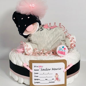 K&ouml;nnte beinhalten: Eine Windeltorte, verziert mit rosa und schwarzem Band, einem rosa Tutu, einer schwarz-wei&szlig;en Polka-Dot-M&uuml;tze und einem rosa Schnuller. Die Torte ist mit einem Schild verziert, auf dem "It's a GIRL!" und "Sailor Marie" steht.