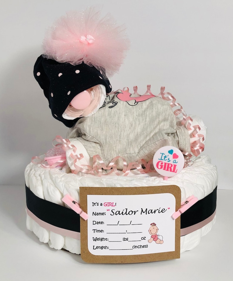K&ouml;nnte beinhalten: Eine Windeltorte, verziert mit rosa und schwarzem Band, einem rosa Tutu, einer schwarz-wei&szlig;en Polka-Dot-M&uuml;tze und einem rosa Schnuller. Die Torte ist mit einem Schild verziert, auf dem "It's a GIRL!" und "Sailor Marie" steht.