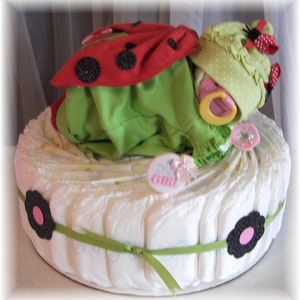 K&ouml;nnte beinhalten: Eine Windeltorte, dekoriert mit einem Marienk&auml;fer-Baby-Outfit, einer gr&uuml;n-wei&szlig; gepunkteten M&uuml;tze und einem gelben Schnuller. Die Torte ist mit einem gr&uuml;nen Band umwickelt und hat an der Seite eine schwarze Blume mit einem rosa Zentrum. Die Torte ist mit "Es ist ein M&auml;dchen!" beschriftet.
