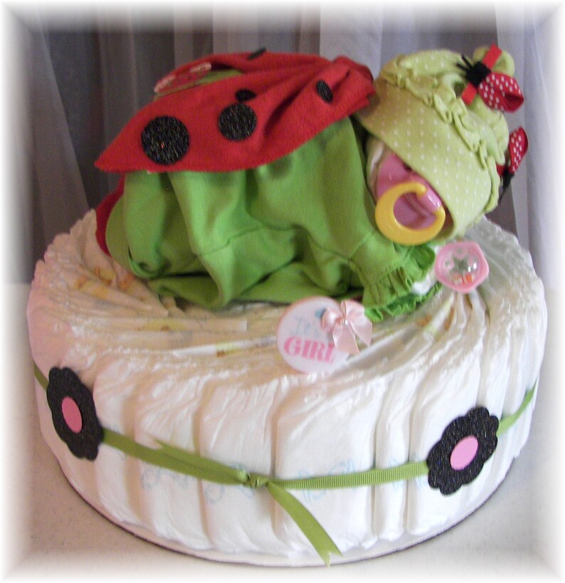 K&ouml;nnte beinhalten: Eine Windeltorte, dekoriert mit einem Marienk&auml;fer-Baby-Outfit, einer gr&uuml;n-wei&szlig; gepunkteten M&uuml;tze und einem gelben Schnuller. Die Torte ist mit einem gr&uuml;nen Band umwickelt und hat an der Seite eine schwarze Blume mit einem rosa Zentrum. Die Torte ist mit "Es ist ein M&auml;dchen!" beschriftet.