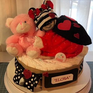 K&ouml;nnte beinhalten: Eine Windeltorte, verziert mit einem rosa Teddyb&auml;r, einem rot-schwarz gestreiften Outfit und einem schwarz-wei&szlig; gepunkteten Band. Die Torte ist mit einem Schild mit der Aufschrift "It's a Girl" und einem Schild mit der Aufschrift "ELORA" verziert.