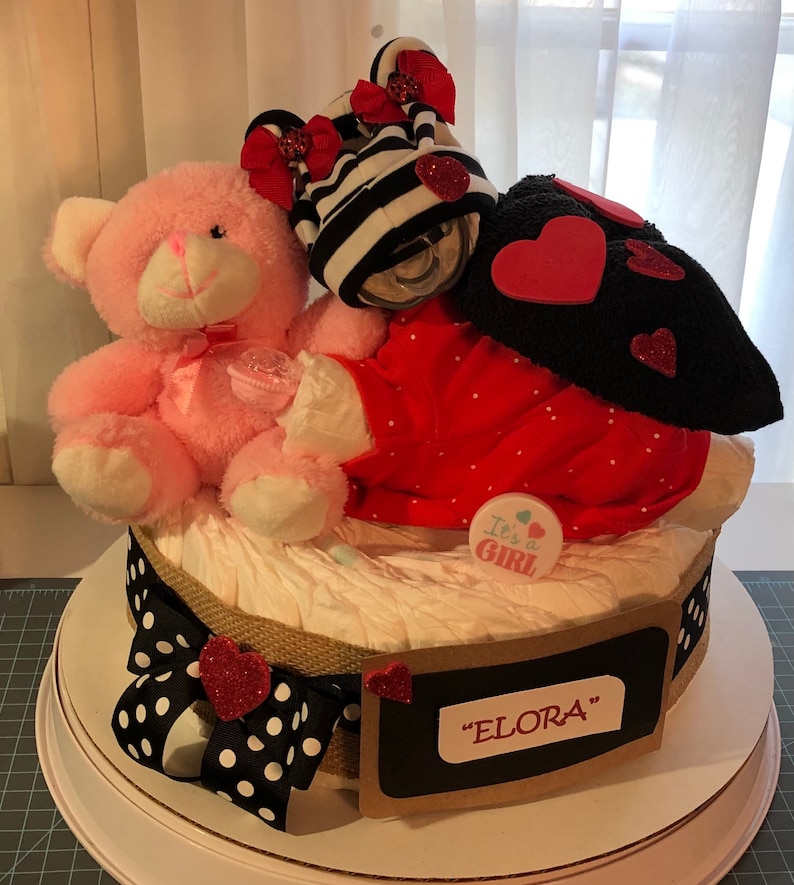 K&ouml;nnte beinhalten: Eine Windeltorte, verziert mit einem rosa Teddyb&auml;r, einem rot-schwarz gestreiften Outfit und einem schwarz-wei&szlig; gepunkteten Band. Die Torte ist mit einem Schild mit der Aufschrift "It's a Girl" und einem Schild mit der Aufschrift "ELORA" verziert.