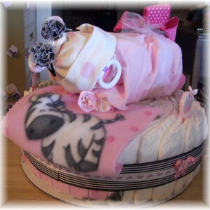 K&ouml;nnte beinhalten: Eine rosa und wei&szlig;e Windeltorte, verziert mit einer Zebra-Decke, einem rosa und wei&szlig;en Tupfen-Strampler und einer rosa Schleife. Die Torte ist mit einem schwarz-wei&szlig;en Band umwickelt.
