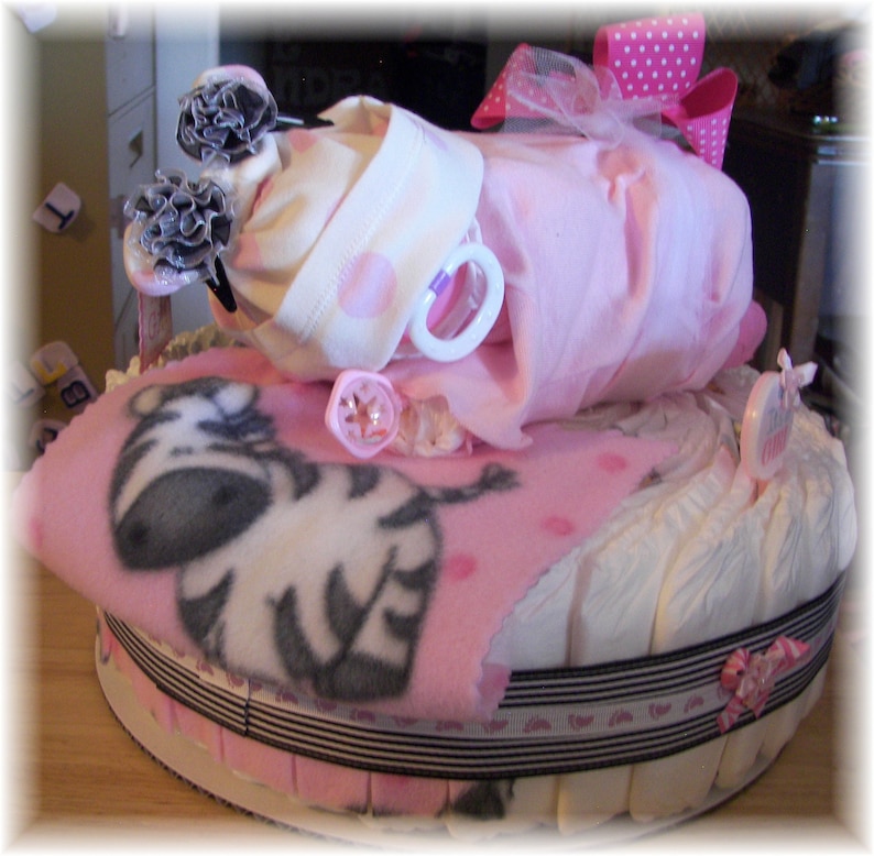 K&ouml;nnte beinhalten: Eine rosa und wei&szlig;e Windeltorte, verziert mit einer Zebra-Decke, einem rosa und wei&szlig;en Tupfen-Strampler und einer rosa Schleife. Die Torte ist mit einem schwarz-wei&szlig;en Band umwickelt.