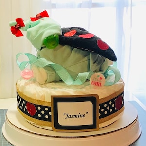 K&ouml;nnte beinhalten: Eine zweist&ouml;ckige Windeltorte, verziert mit schwarz-wei&szlig;em Polka-Dot-Band, roten Herzen und einer gr&uuml;n-wei&szlig;en Baby-M&uuml;tze. Die Torte ist mit einem schwarzen und roten Marienk&auml;fer-Pl&uuml;schtier und einem Schild mit der Aufschrift "It's a Girl!" gekr&ouml;nt. Die untere Schicht hat ein Schild mit der Aufschrift "Jasmine".