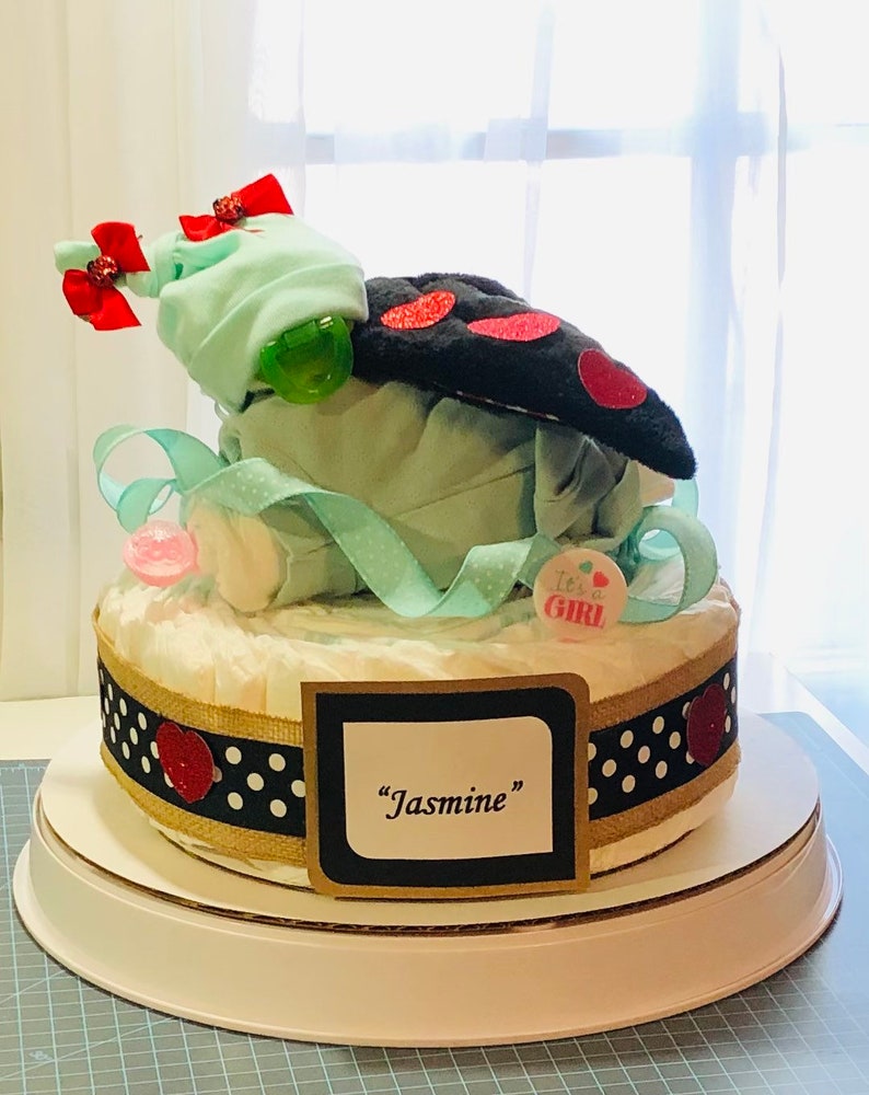 K&ouml;nnte beinhalten: Eine zweist&ouml;ckige Windeltorte, verziert mit schwarz-wei&szlig;em Polka-Dot-Band, roten Herzen und einer gr&uuml;n-wei&szlig;en Baby-M&uuml;tze. Die Torte ist mit einem schwarzen und roten Marienk&auml;fer-Pl&uuml;schtier und einem Schild mit der Aufschrift "It's a Girl!" gekr&ouml;nt. Die untere Schicht hat ein Schild mit der Aufschrift "Jasmine".