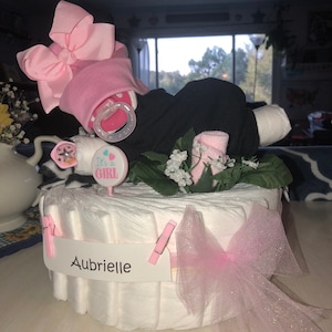 K&ouml;nnte beinhalten: Eine Windeltorte, verziert mit einer rosa Schleife, einem Schnuller und einem Schild mit der Aufschrift "It's a Girl". Die Torte ist au&szlig;erdem mit einer rosa T&uuml;llschleife und einem wei&szlig;en Banner mit der Aufschrift "Aubrielle" verziert.