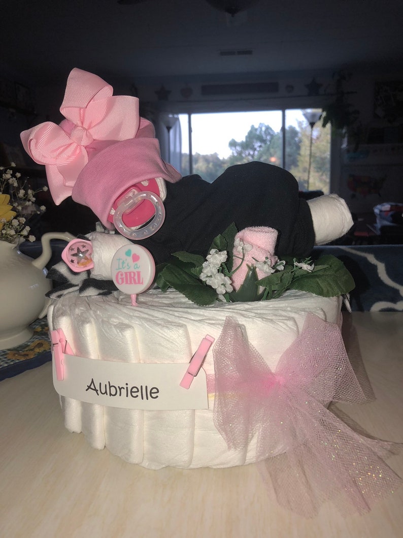 K&ouml;nnte beinhalten: Eine Windeltorte, verziert mit einer rosa Schleife, einem Schnuller und einem Schild mit der Aufschrift "It's a Girl". Die Torte ist au&szlig;erdem mit einer rosa T&uuml;llschleife und einem wei&szlig;en Banner mit der Aufschrift "Aubrielle" verziert.