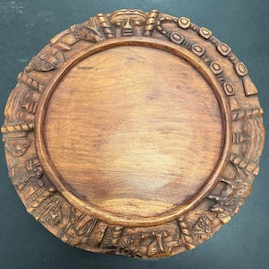 Puede incluir: Una bandeja decorativa redonda de madera con intrincadas tallas alrededor del borde. Las tallas representan figuras humanas y patrones geométricos. La bandeja es de color marrón medio cálido y tiene un centro liso y plano.