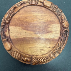 Puede incluir: Una bandeja redonda de madera con un borde tallado. El borde presenta varios diseños tallados, incluyendo caras, botellas y otras formas abstractas. La bandeja tiene un acabado de veta de madera natural y probablemente está hecha a mano.