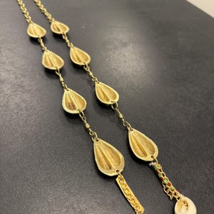 Può includere: Orecchini dorati con catena e pendenti a forma di goccia. Gli orecchini hanno una superficie strutturata e una piccola catena con perline colorate e una conchiglia bianca all'estremità. Su una superficie scura.