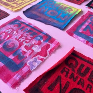 May include: A selection of vibrant fabric squares, each bearing the words "QUEER ANARCHY NOW". The squares are in shades of red, yellow, blue, and pink, with the text in contrasting hues. The fabric edges are raw and unfinished, giving a handmade feel.