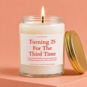 May include: A lit soy wax candle in a clear glass jar with a gold metal lid. The white label features red text reading LA CANDELA, Turning 25 For The Third Time, and smaller text describing scents of denial, birthday cake, and anti-aging cream.