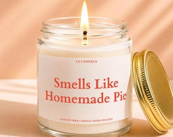 Może przedstawiać: Świeca w przezroczystym szklanym słoiku z białą etykietą z napisem LA CANDELA oraz Smells Like Homemade Pie czerwonymi literami. Świeca zawiera biały wosk, ma zapalony knot i oddzielną złotą metalową pokrywkę.