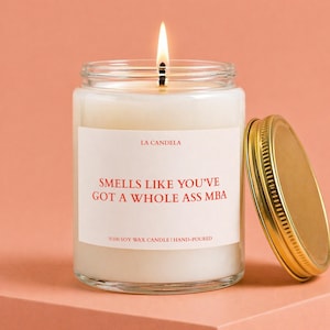 May include: A lit soy wax candle in a clear glass jar with a gold lid. The label reads "SMELLS LIKE YOU'VE GOT A WHOLE ASS MBA" in red text. The candle is hand-poured. The background is a peach color.