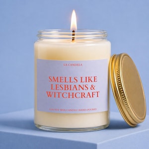 May include: A lit soy wax candle in a glass jar with a gold lid. The label reads "SMELLS LIKE LESBIANS & WITCHCRAFT" in red text. The candle is hand-poured and has the text "LA CANDELA" at the top of the label.