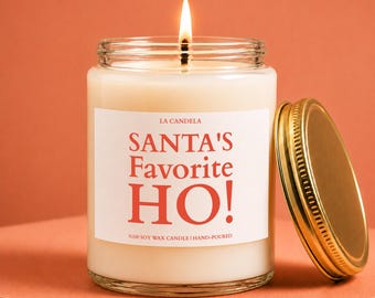 Może przedstawiać: Świeca w przezroczystym szklanym słoiku z białą etykietą z czerwonym napisem LA CANDELA SANTA'S Favorite HO! %100 SOY WAX CANDLE | HAND-POURED. Świeca ma zapalony knot, a obok słoika znajduje się złota metalowa pokrywka.