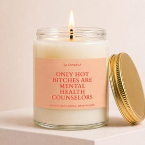 May include: A lit soy wax candle in a clear glass jar with a gold lid. The label reads "ONLY HOT BITCHES ARE MENTAL HEALTH COUNSELORS" in red text. The candle is hand-poured and from La Candela.