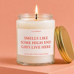 May include: A clear glass jar candle with a white wax fill and a lit wick. The label features the red text LA CANDELA and SMELLS LIKE SOME HIGH END GAYS LIVE HERE, with %100 SOY WAX CANDLE | HAND-POURED at the bottom. A gold metal lid sits to the right.