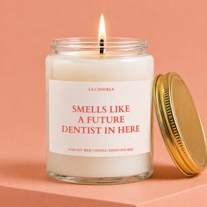 May include: A burning candle in a clear glass jar with a gold lid. The label says "SMELLS LIKE A FUTURE DENTIST IN HERE." The candle is labelled "LA CANDELA" and is made of 100% soy wax.