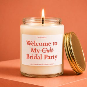 A white candle with a lit flame, labeled "Welcome to My-Gult Bridal Party" and "100% Soy Wax Candle Hand-Poured", is placed on a pink surface.