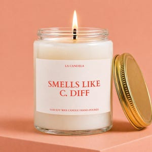 May include: A lit soy wax candle in a clear glass jar with a gold lid. The label reads "Smells Like C. Diff" in red text. The candle is hand-poured and made with 100% soy wax.