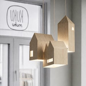 Puede incluir: Tres l&aacute;mparas colgantes de madera con forma de casa que cuelgan del techo. Cada l&aacute;mpara tiene forma rectangular con un tejado a dos aguas y una peque&ntilde;a ventana rectangular. Las l&aacute;mparas son de madera clara y est&aacute;n iluminadas desde dentro.