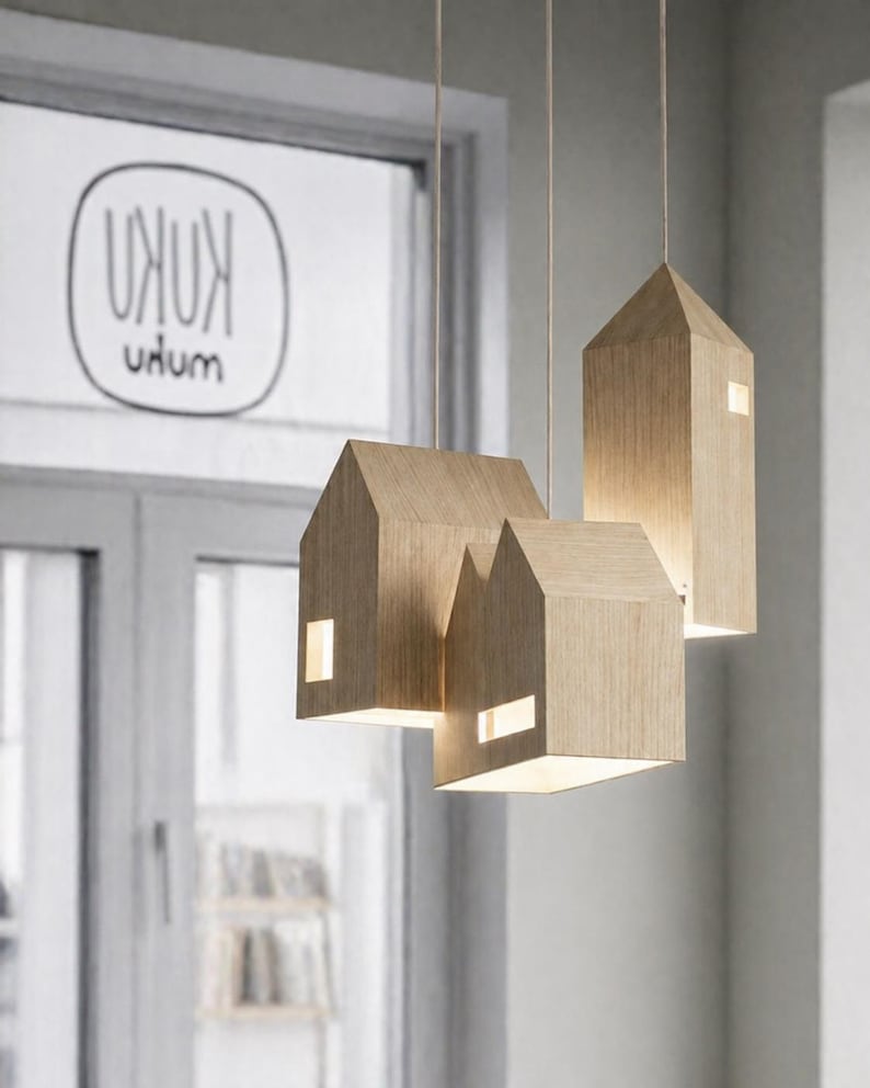 Puede incluir: Tres l&aacute;mparas colgantes de madera con forma de casa que cuelgan del techo. Cada l&aacute;mpara tiene forma rectangular con un tejado a dos aguas y una peque&ntilde;a ventana rectangular. Las l&aacute;mparas son de madera clara y est&aacute;n iluminadas desde dentro.