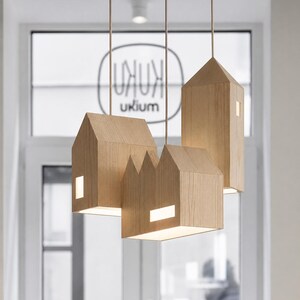 Puede incluir: Un conjunto de l&aacute;mparas colgantes de madera con forma de casa. Cada l&aacute;mpara tiene un tama&ntilde;o y forma diferentes, con ventanas rectangulares que emiten un brillo c&aacute;lido. Las luces est&aacute;n suspendidas del techo por finos cordones, creando un dise&ntilde;o moderno.