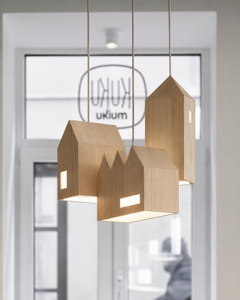 Puede incluir: Un conjunto de l&aacute;mparas colgantes de madera con forma de casa. Cada l&aacute;mpara tiene un tama&ntilde;o y forma diferentes, con ventanas rectangulares que emiten un brillo c&aacute;lido. Las luces est&aacute;n suspendidas del techo por finos cordones, creando un dise&ntilde;o moderno.