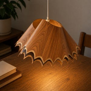 May include: A wooden pendant light fixture with a scalloped edge design. The light has a warm wood grain pattern and is suspended from a white cord. The fixture is hanging over a wooden table.