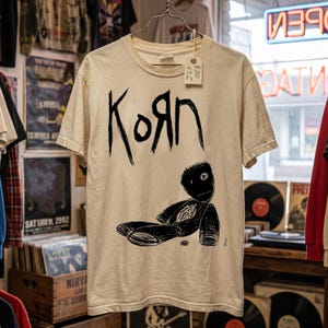 May include: A cream-colored short-sleeve t-shirt featuring a black graphic print of the band name Korn in a stylized font above a drawing of a stitched doll. A small white price tag is attached to the collar with a string.